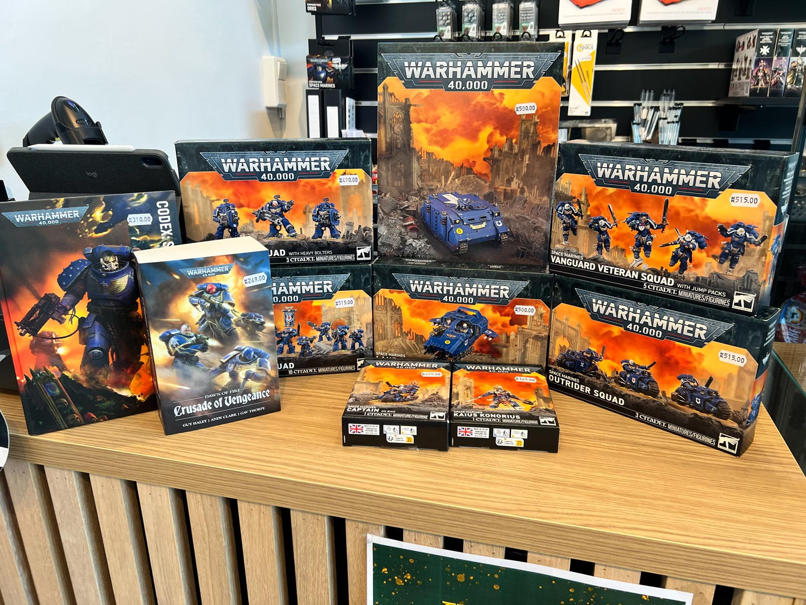 En utstillingshylle i tre fylt med ulike Warhammer 40 000-bokssett og en bok, med Space Marine-figurer og kjøretøy.